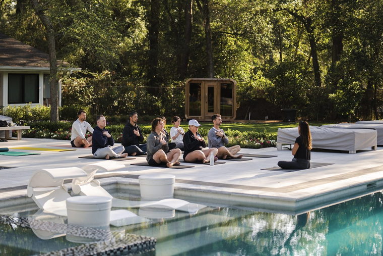 A seated breathwork session on mats by the pool in early light, hands resting on chests, the sauna behind.