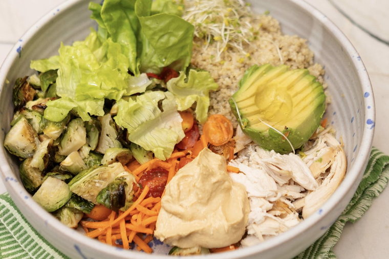 A lunch bowl of quinoa, roasted brussels sprouts, shredded chicken, avocado, carrot and hummus.