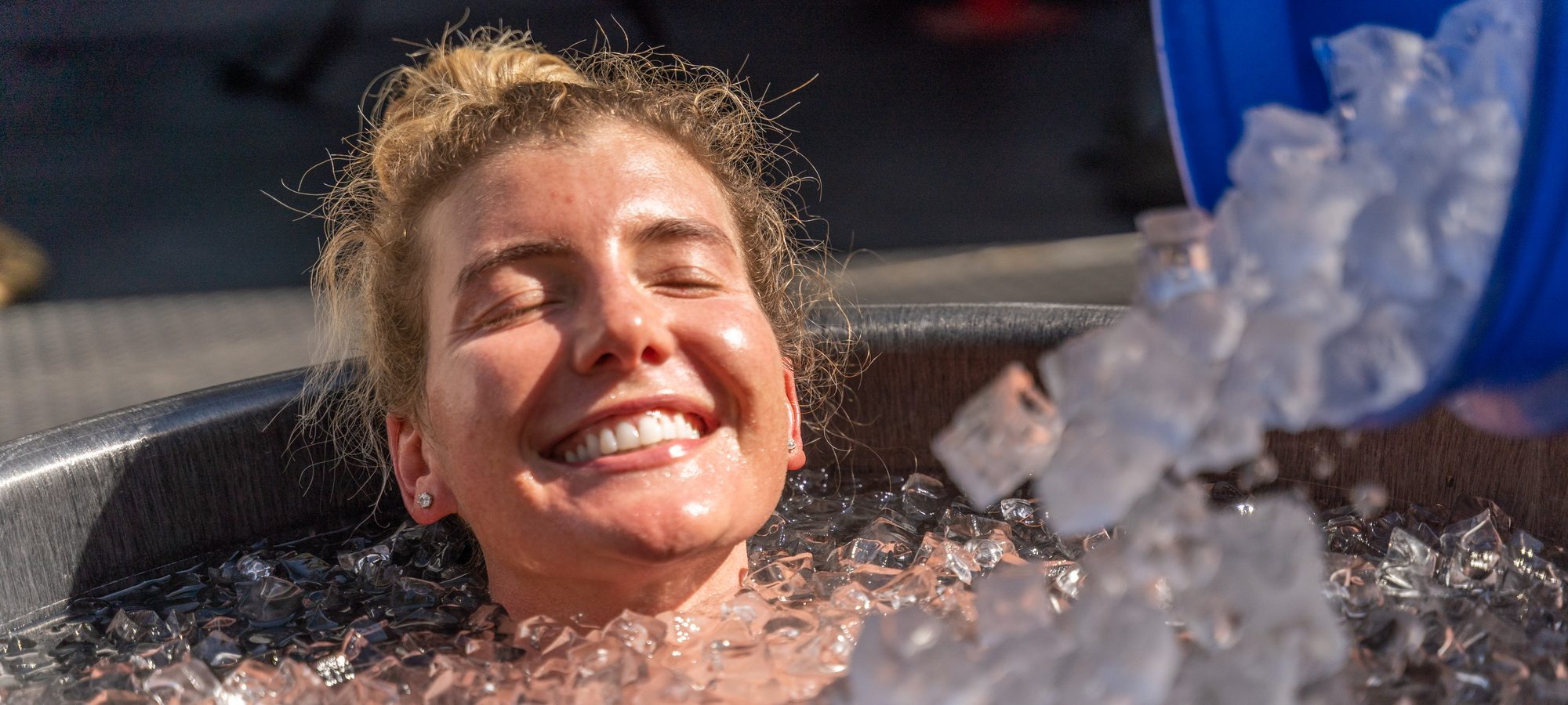 Fresh ice being poured into a plunge tub as an attendee sits in it, smiling.
